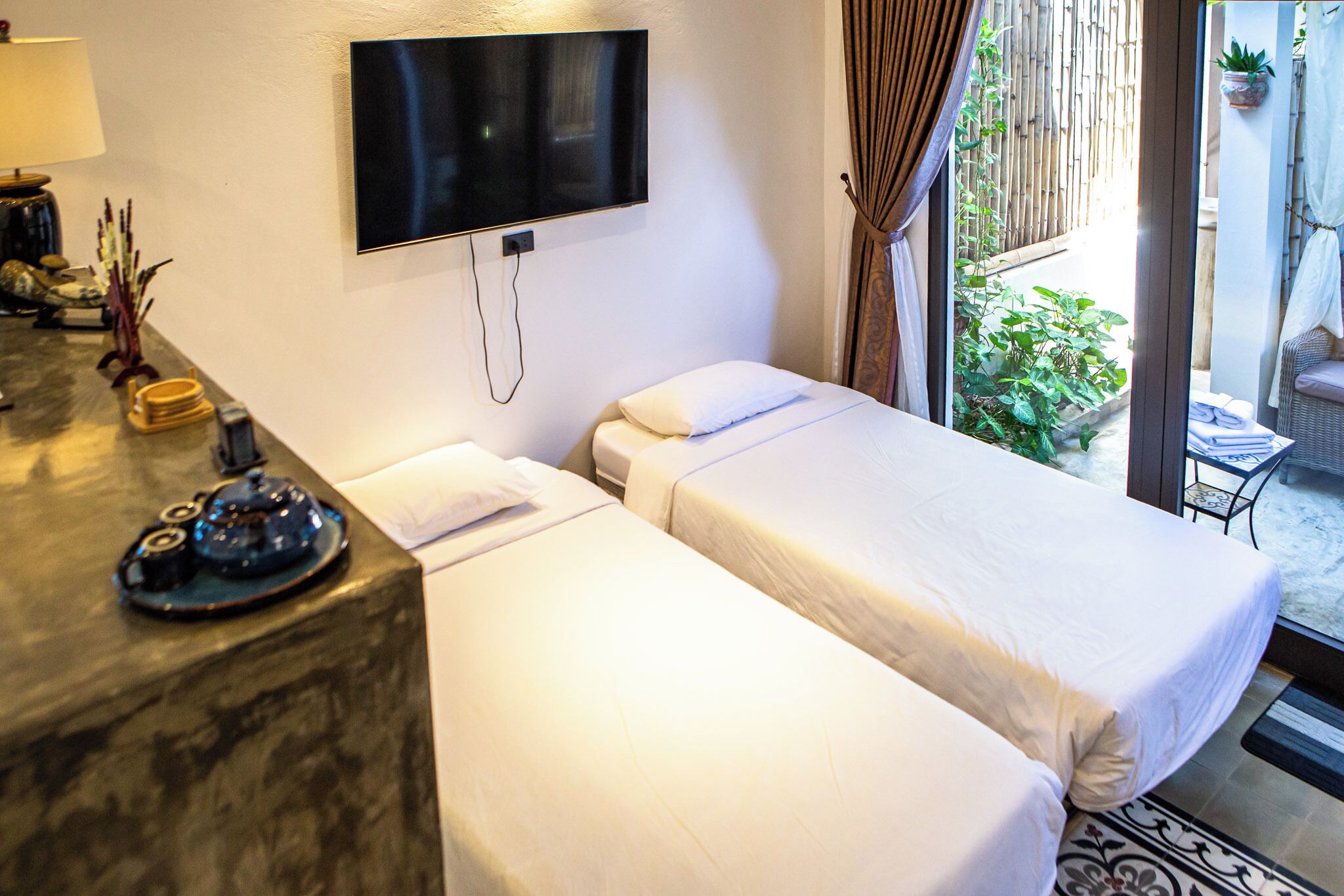 A hotel room with two beds, a TV mounted on the wall, a small table with a lamp and decorative items, and a view of the outdoor area.