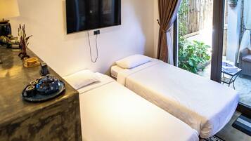 A hotel room with two beds, a TV mounted on the wall, a small table with a lamp and decorative items, and a view of the outdoor area.