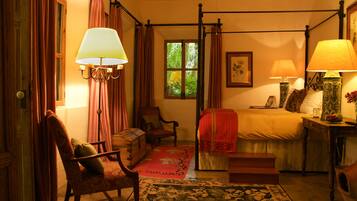 A traditional bedroom with a four-poster bed, a sitting area with a chair and a small table, and a window with curtains.