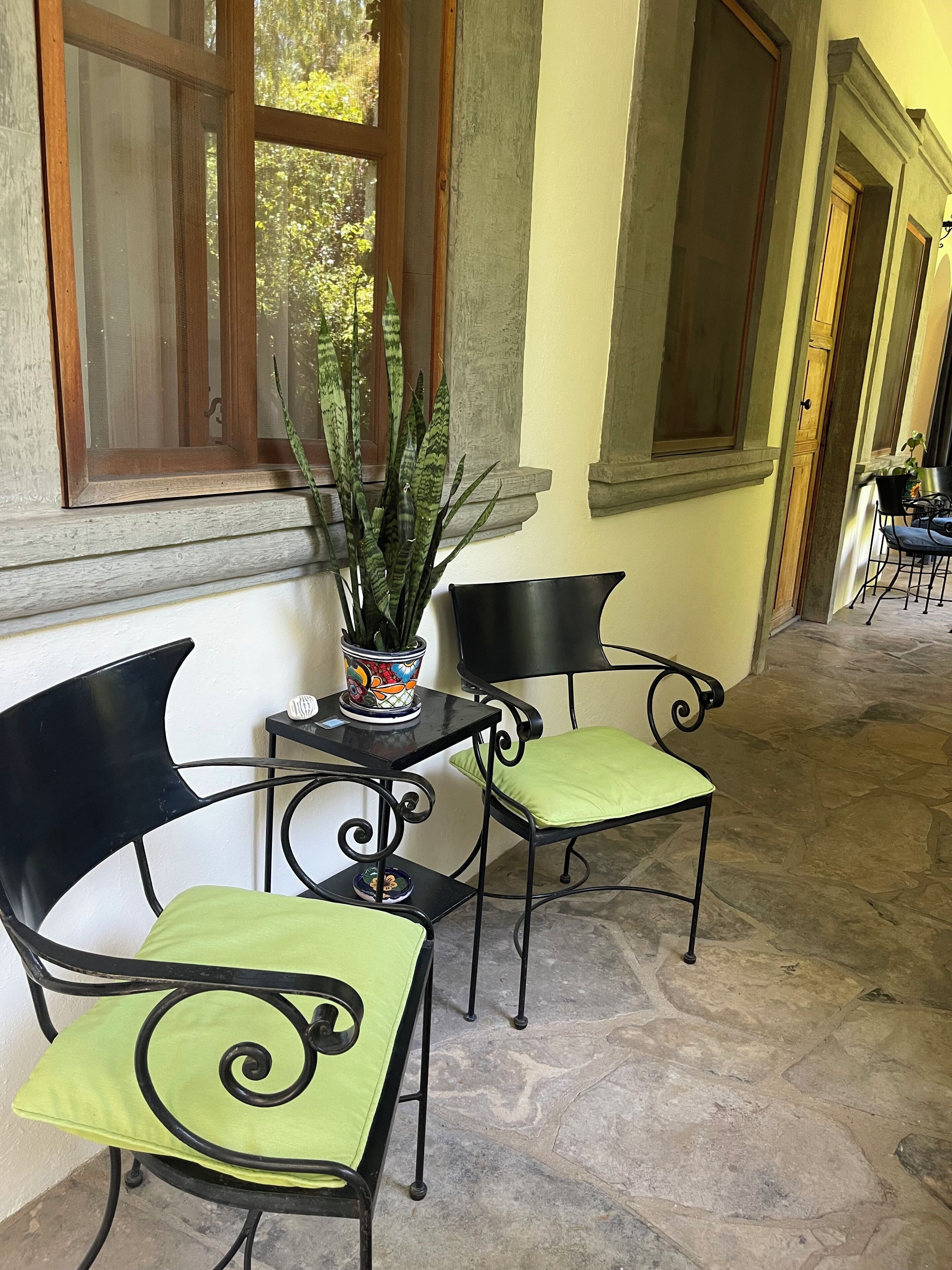 Una habitación con piso de piedra, dos sillas de metal negro con cojines verdes, una mesita con planta en maceta y una ventana con vistas a árboles.