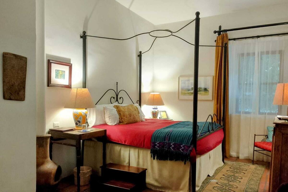 Ein Bett mit Baldachin, mit roter Bettdecke, zwei Nachttische mit Lampen, ein Holzstuhl, ein kleiner Teppich und ein Fenster mit Vorhängen.