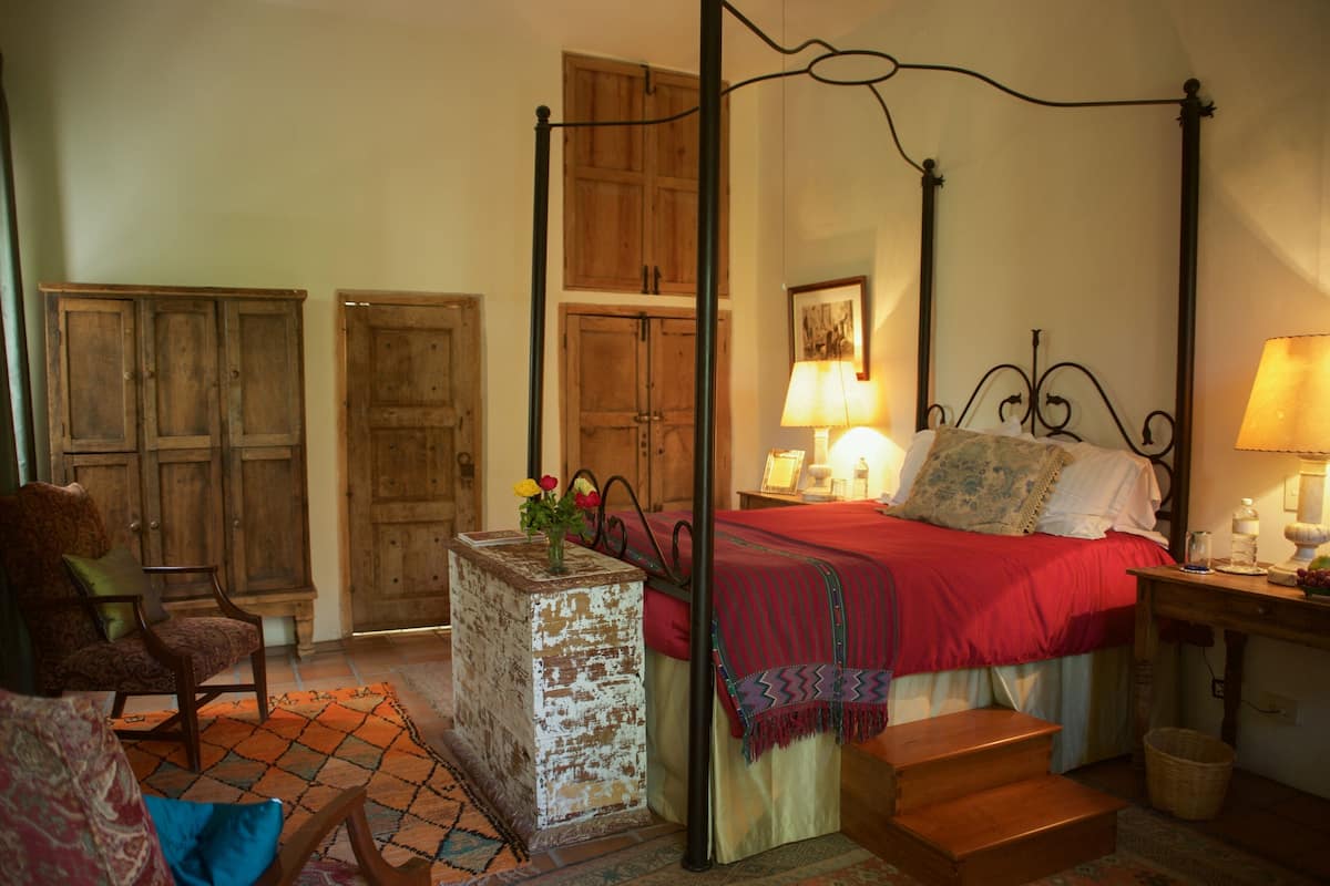 Ein Bett mit Baldachin, mit roter Bettdecke, ein Nachttisch aus Holz mit einer Lampe, ein Stuhl und ein kleiner Tisch mit einem Korb.