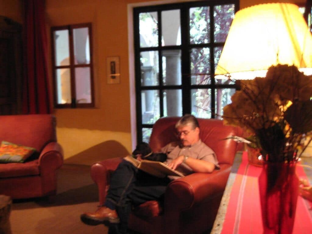 Un hombre sentado en un sillón de cuero leyendo un libro en una habitación cálidamente iluminada, con un ventanal grande y otro sillón rojo.