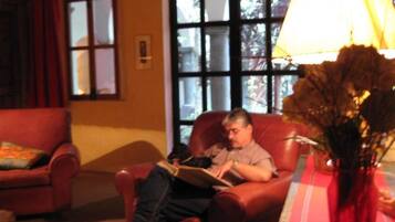 Un hombre sentado en un sillón de cuero leyendo un libro en una habitación cálidamente iluminada, con un ventanal grande y otro sillón rojo.