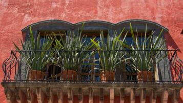 Un balcón con macetas de terracota con plantas verdes, barandilla de hierro forjado y una pared roja.