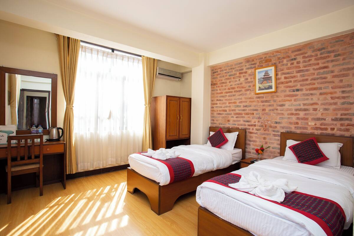 Habitación de hotel con dos camas, un escritorio con silla, un armario y una pared de ladrillo con un cuadro.