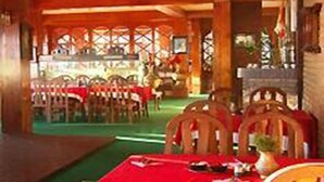 A restaurant with wooden tables and chairs, a green carpet, and a red tablecloth. - Niva Niwa Lodge (Baluwapati Deupur)