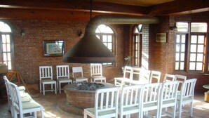 A rustic interior with a stone fireplace surrounded by white chairs, brick walls, and large windows. - Niva Niwa Lodge (Baluwapati Deupur)