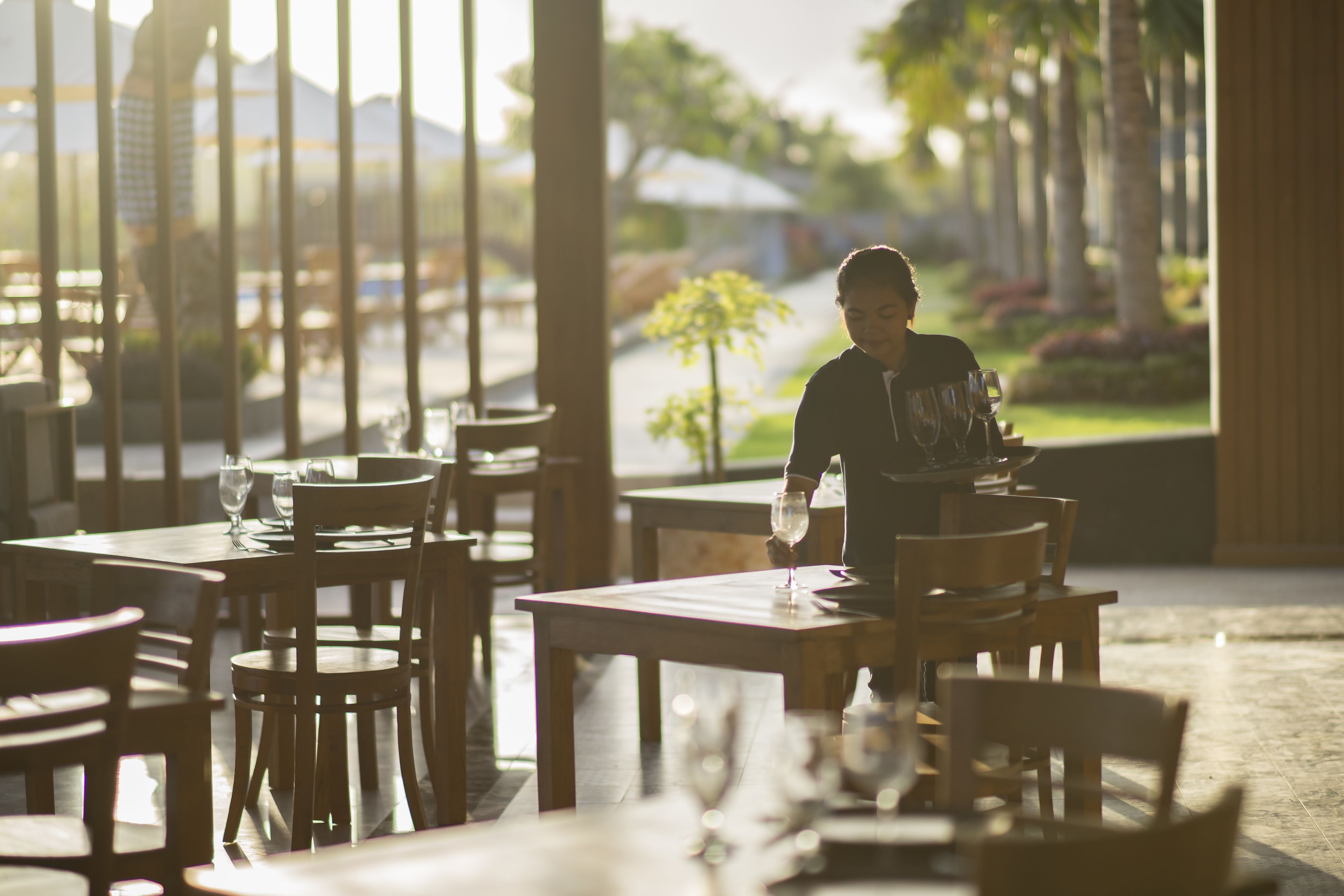 A restaurant with wooden tables and chairs, a waiter serving a customer, and a view of palm trees and a street in the background.