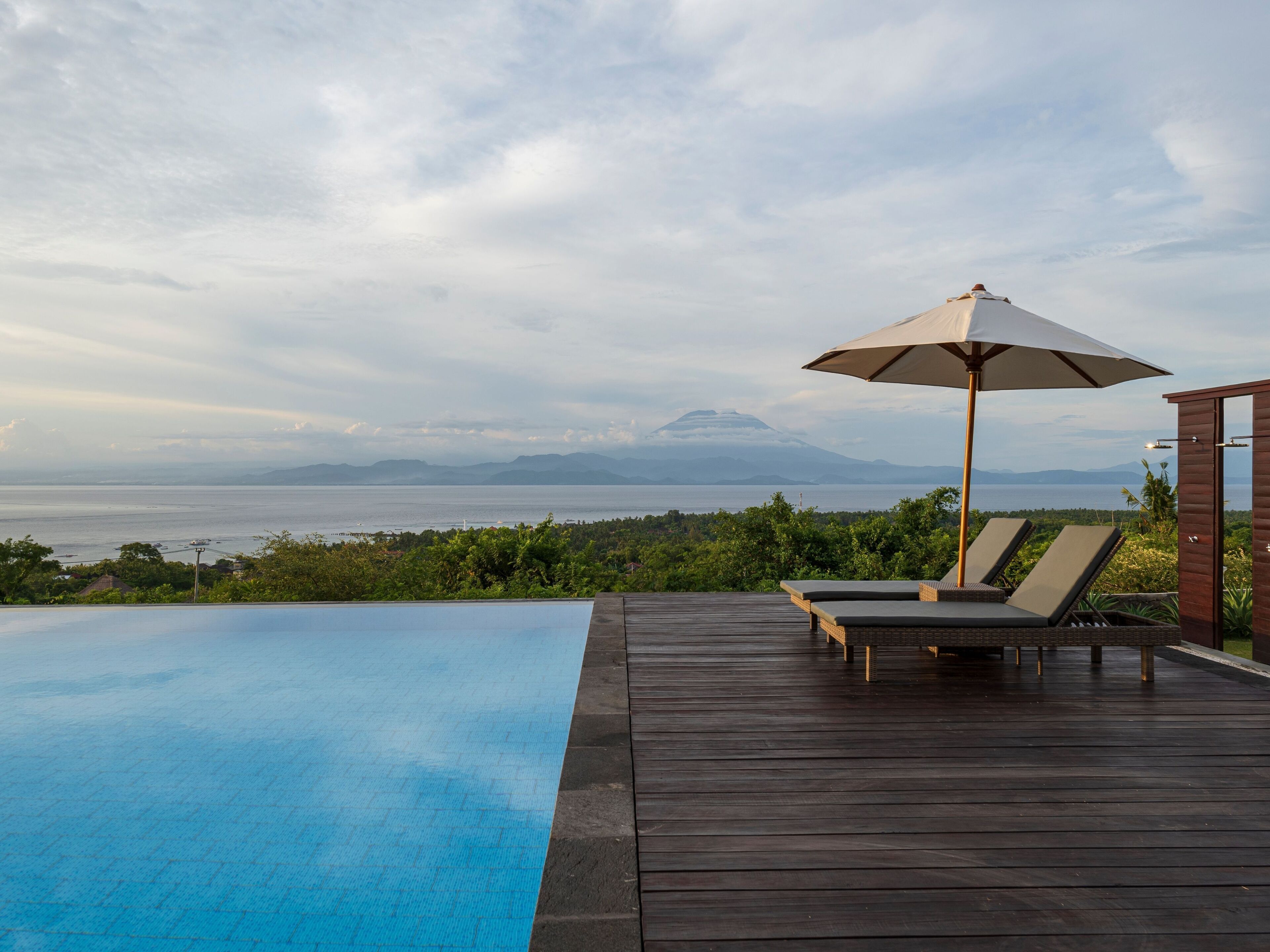 A wooden deck with a pool, lounge chairs, and an umbrella overlooking a body of water and distant mountains.