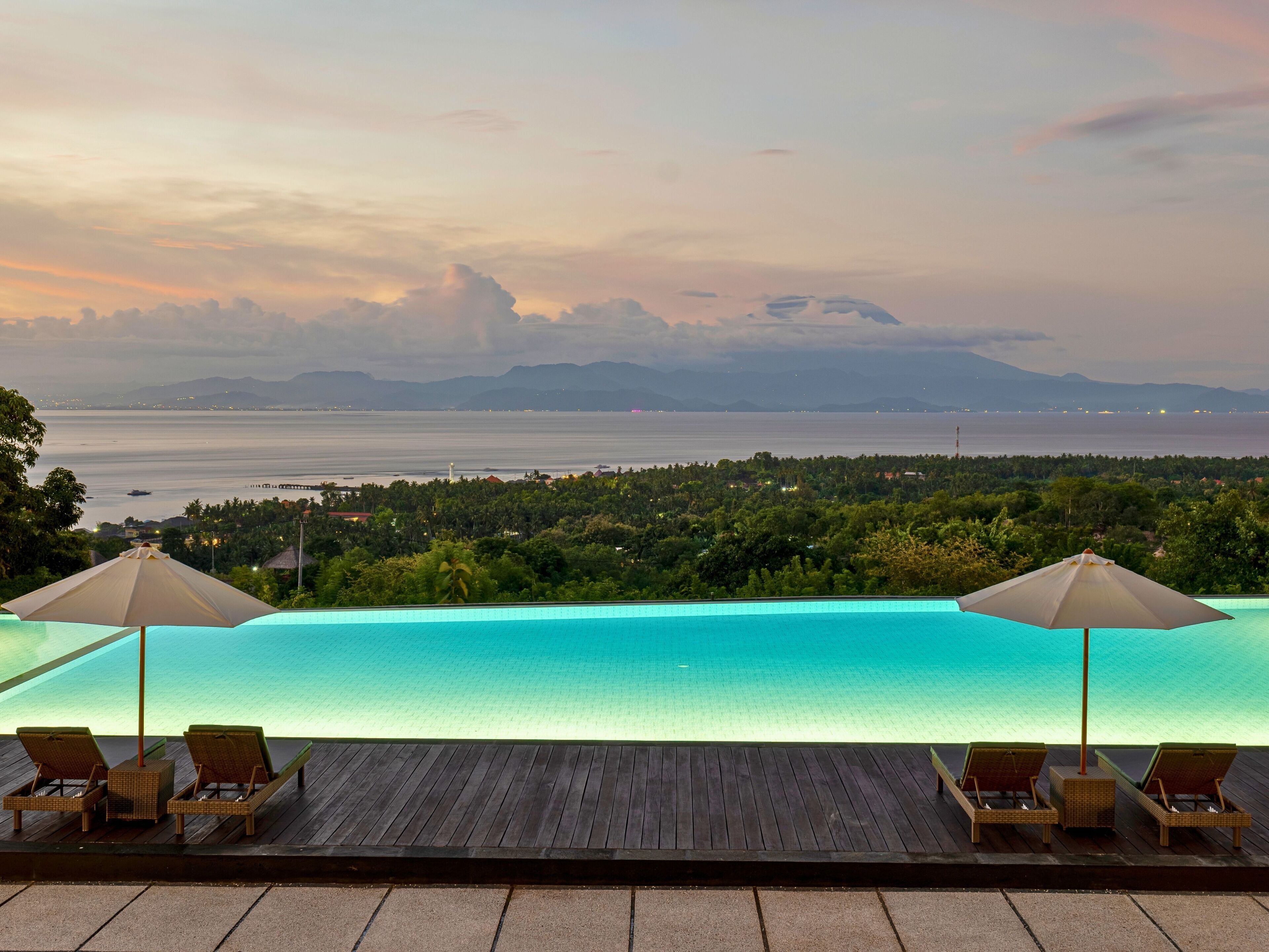 A poolside view with lounge chairs and umbrellas overlooking a body of water and a distant landscape.