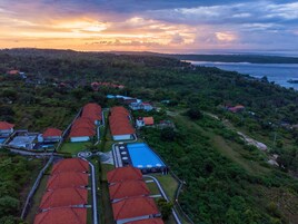 An aerial view of a resort with multiple red-roofed buildings, a swimming pool, and a view of the sea and hills.