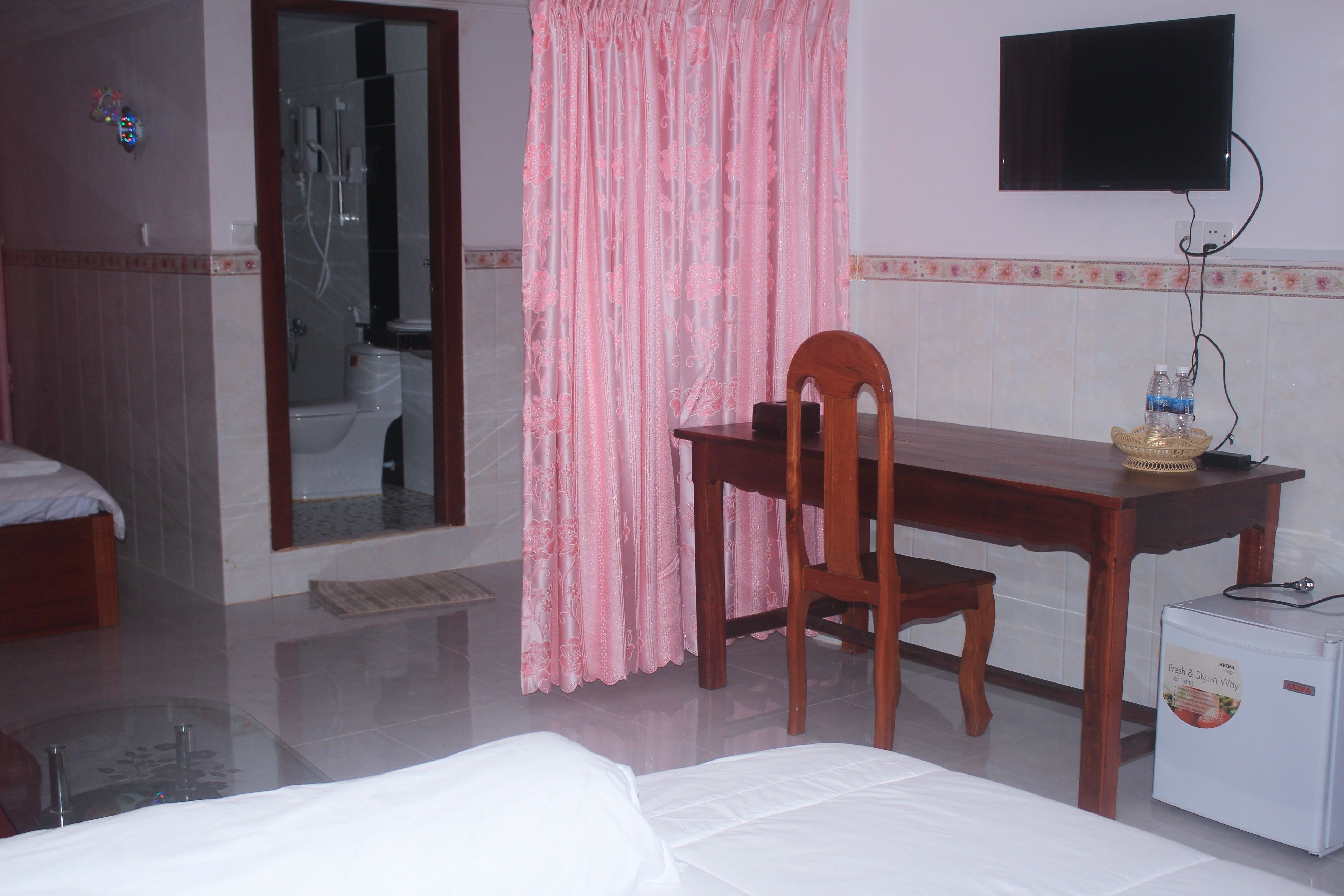 Habitación de hotel con cama, escritorio de madera, televisor, refrigerador y baño visible a través de una puerta abierta.