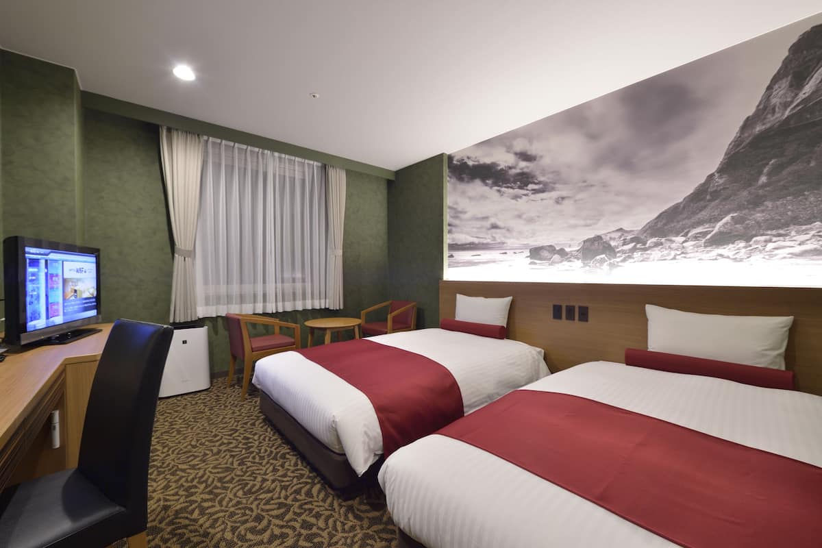 A hotel room with two beds, a desk with a computer, a chair, a small table, and a large window with curtains.