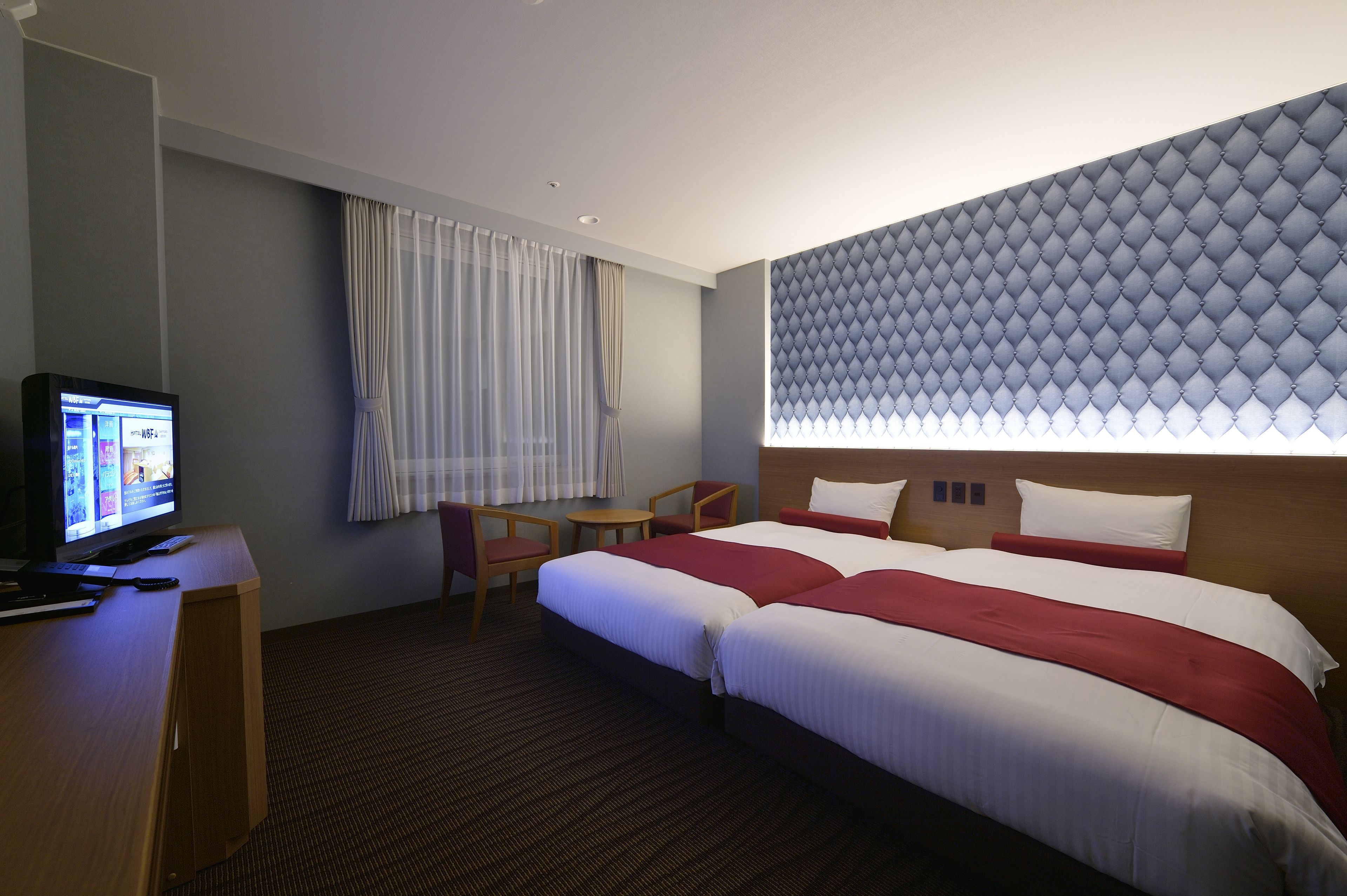 A hotel room with a large bed, a desk with a computer, a small table, and a window with curtains.