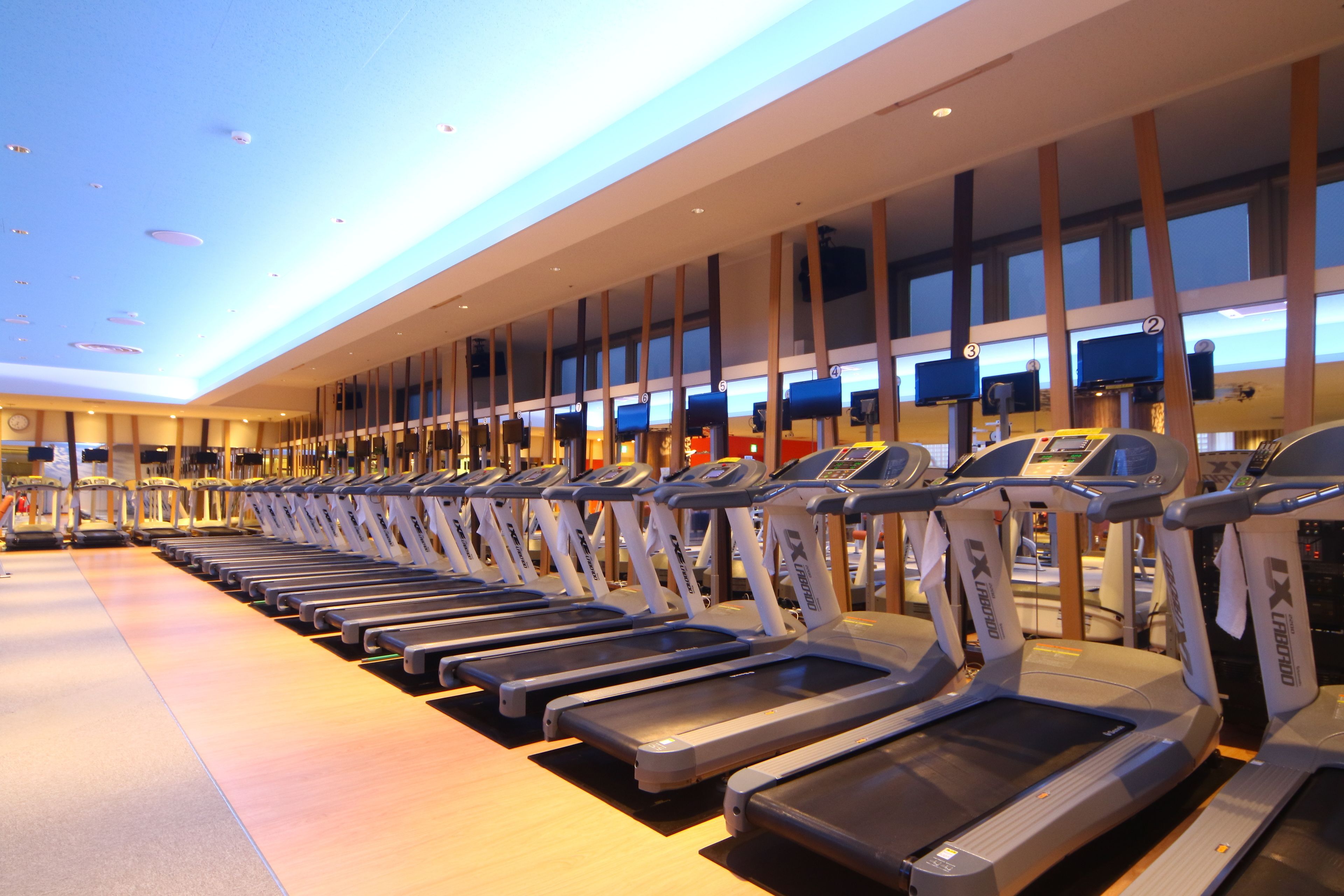 A row of treadmills in a gym with a modern design, large windows, and a ceiling with recessed lighting.