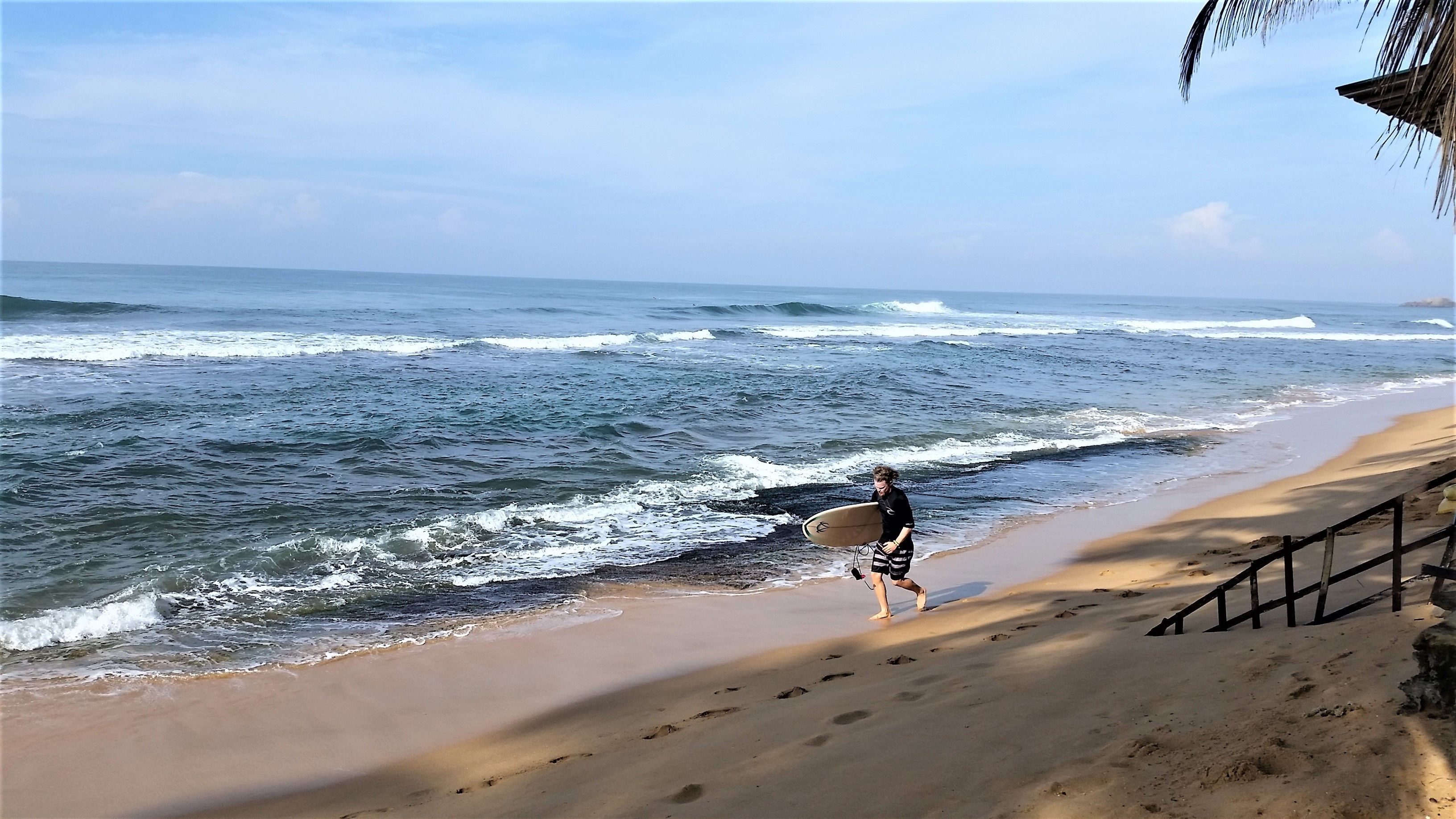 Una playa con olas, una persona cargando una tabla de surf y una valla de madera.