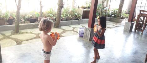 Dos niños jugando con un juguete en un espacio exterior cubierto, con suelo de piedra y plantas en macetas.