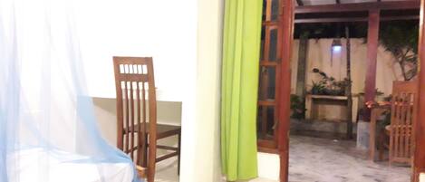 A room with a bed, a chair, and a doorway leading to an outdoor area with plants and a table.