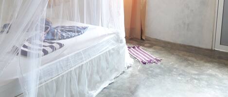 Una habitación con una cama cubierta por una mosquitera blanca, suelo de hormigón y una ventana con cortinas.