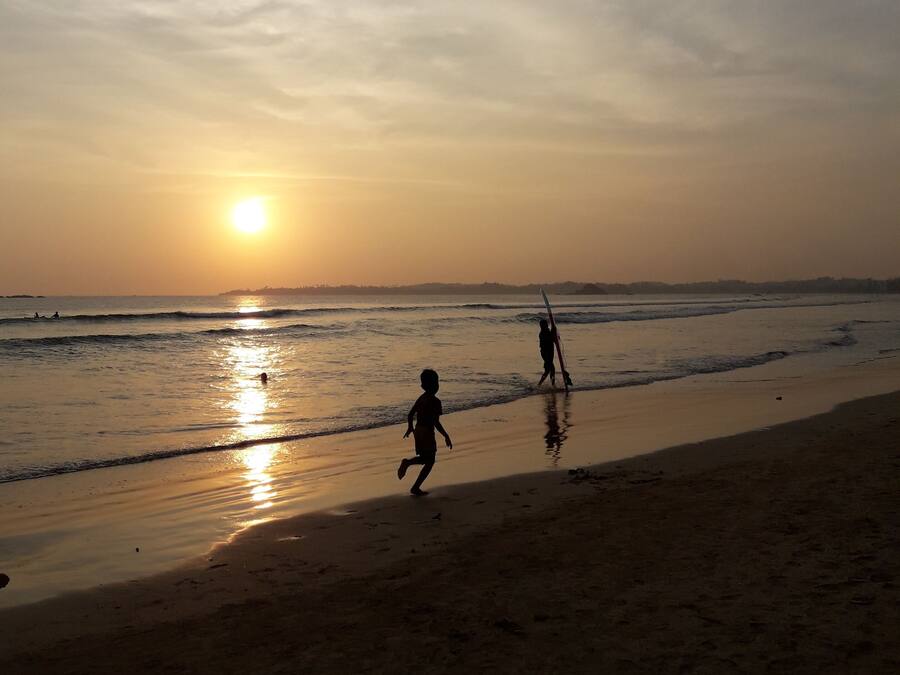 A beach scene with two people walking along the shore, one carrying a surfboard.