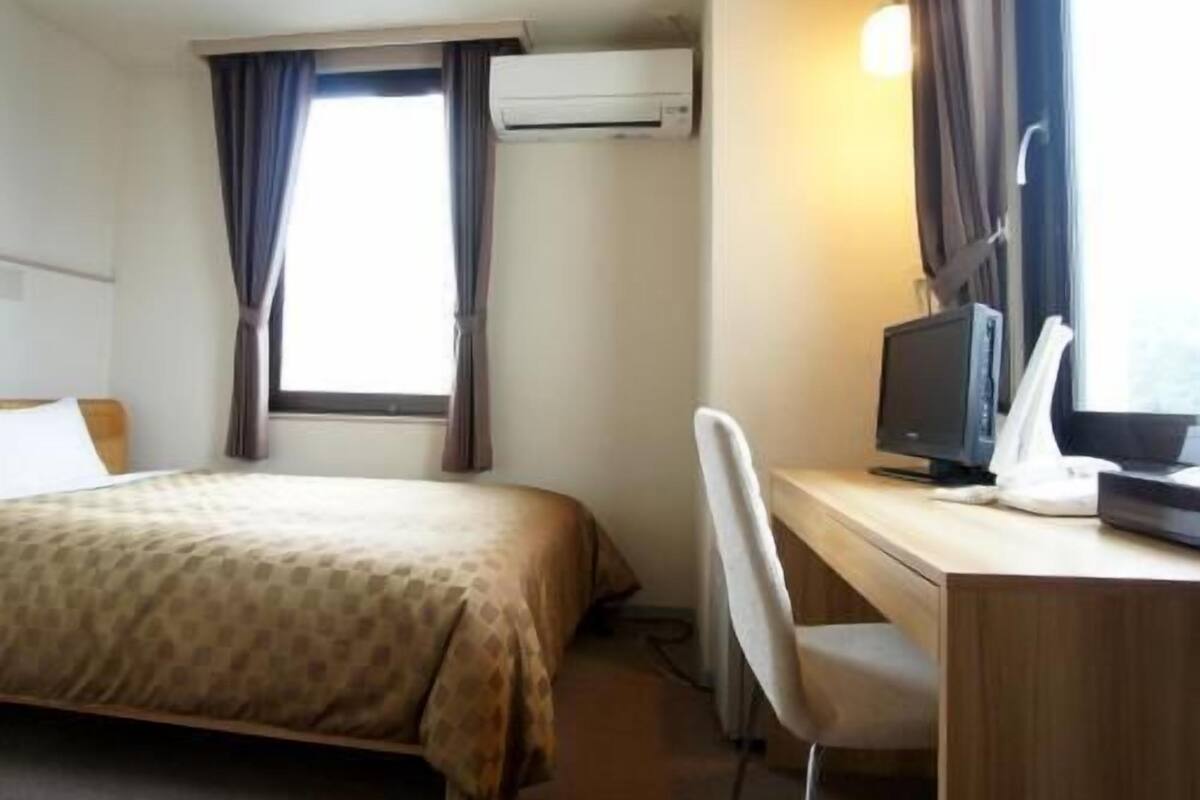 Een hotelkamer met een bed, een bureau met een computer, een airconditioner en een raam met gordijnen.