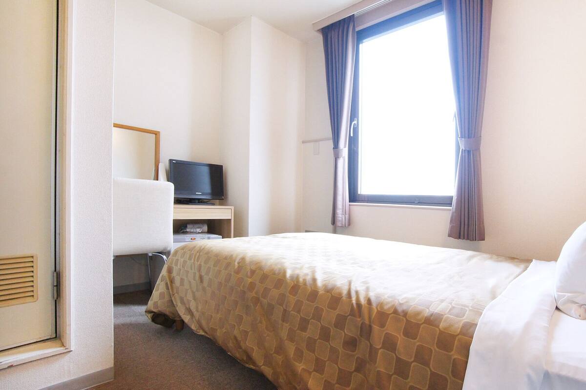 Een hotelkamer met een bed, een bureau met een televisie, een raam met gordijnen en een wandradiator.