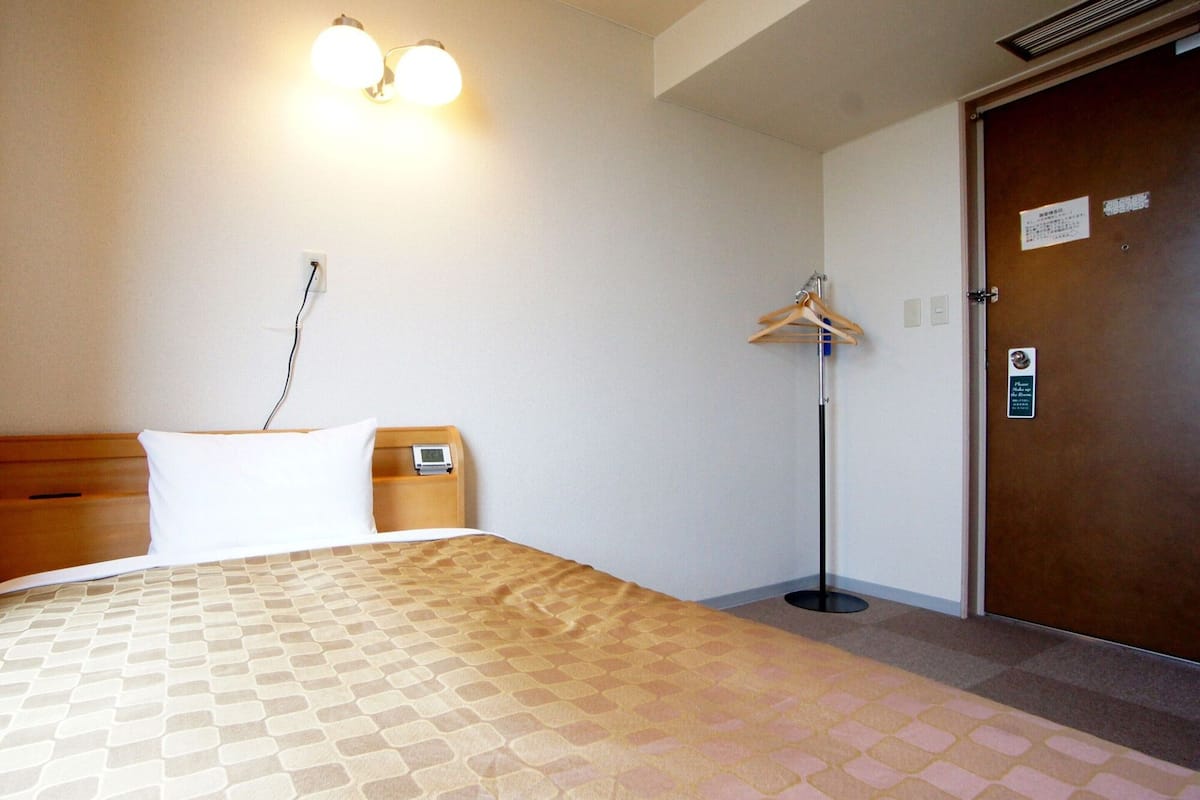 Een hotelkamer met een bed, een nachtkastje met een klok, een kapstok en een deur met een bordje.