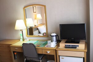 Quarto de hotel com escrivaninha, cadeira, televisão, cafeteira, espelho e um pequeno armário.