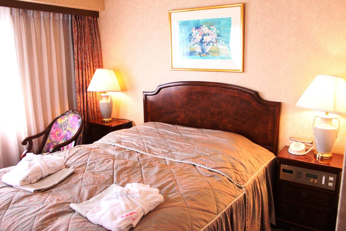 A hotel room with a bed, two bedside lamps, a telephone, a painting on the wall, and a chair with a colorful cushion.