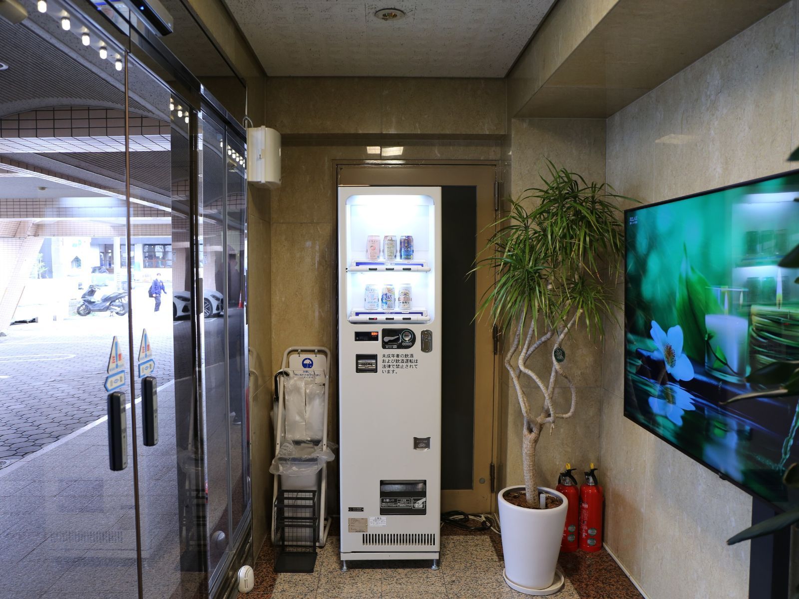 A vending machine, a trash bin, a potted plant, and a fire extinguisher in a hallway.