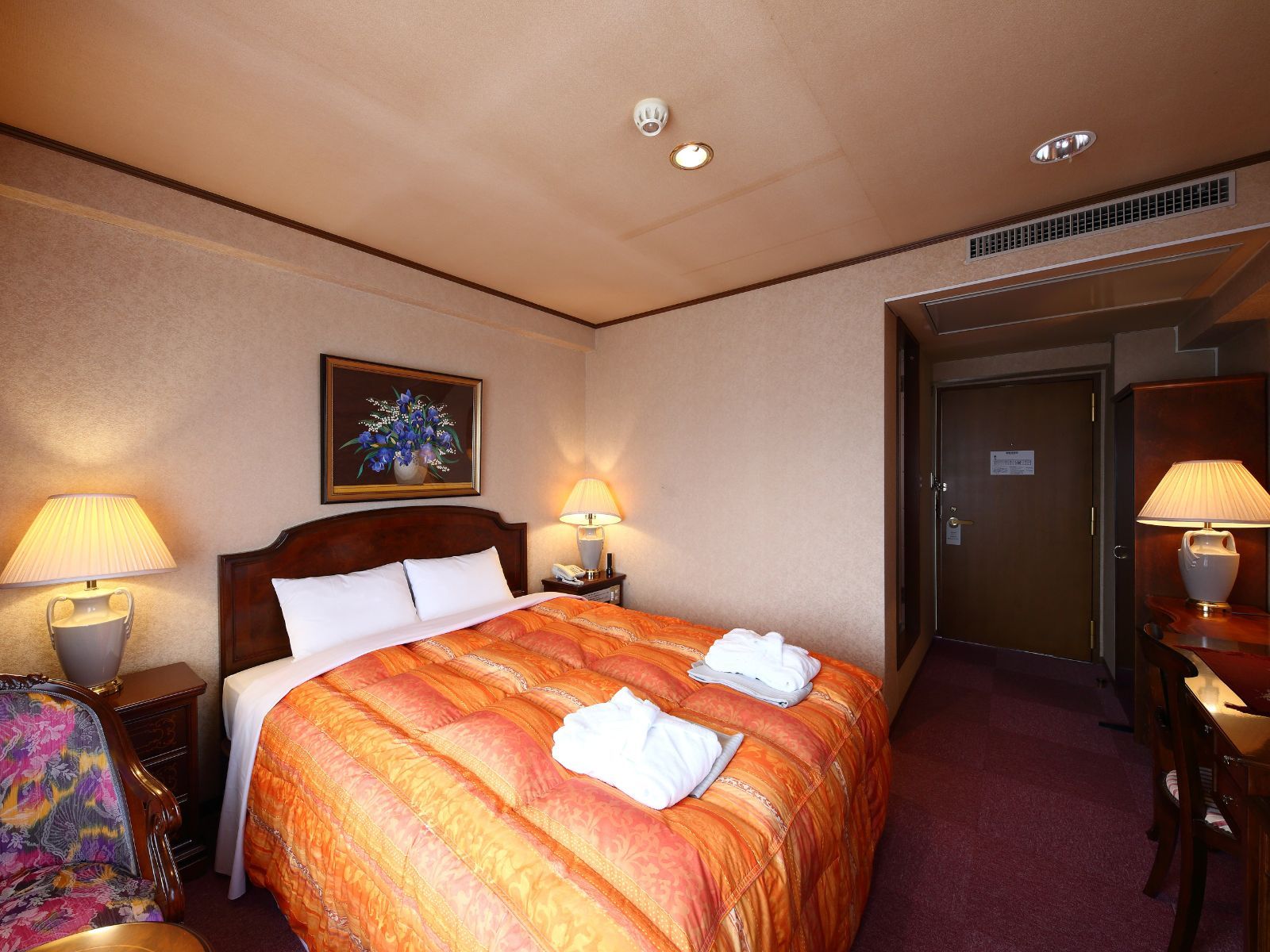 Una camera d'albergo con un letto grande, due abat-jour, un quadro appeso al muro, un ingresso al bagno e una scrivania con una lampada.