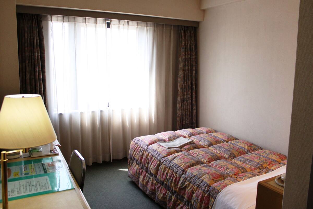 A hotel room with a bed, a desk with a lamp, and a window with curtains.