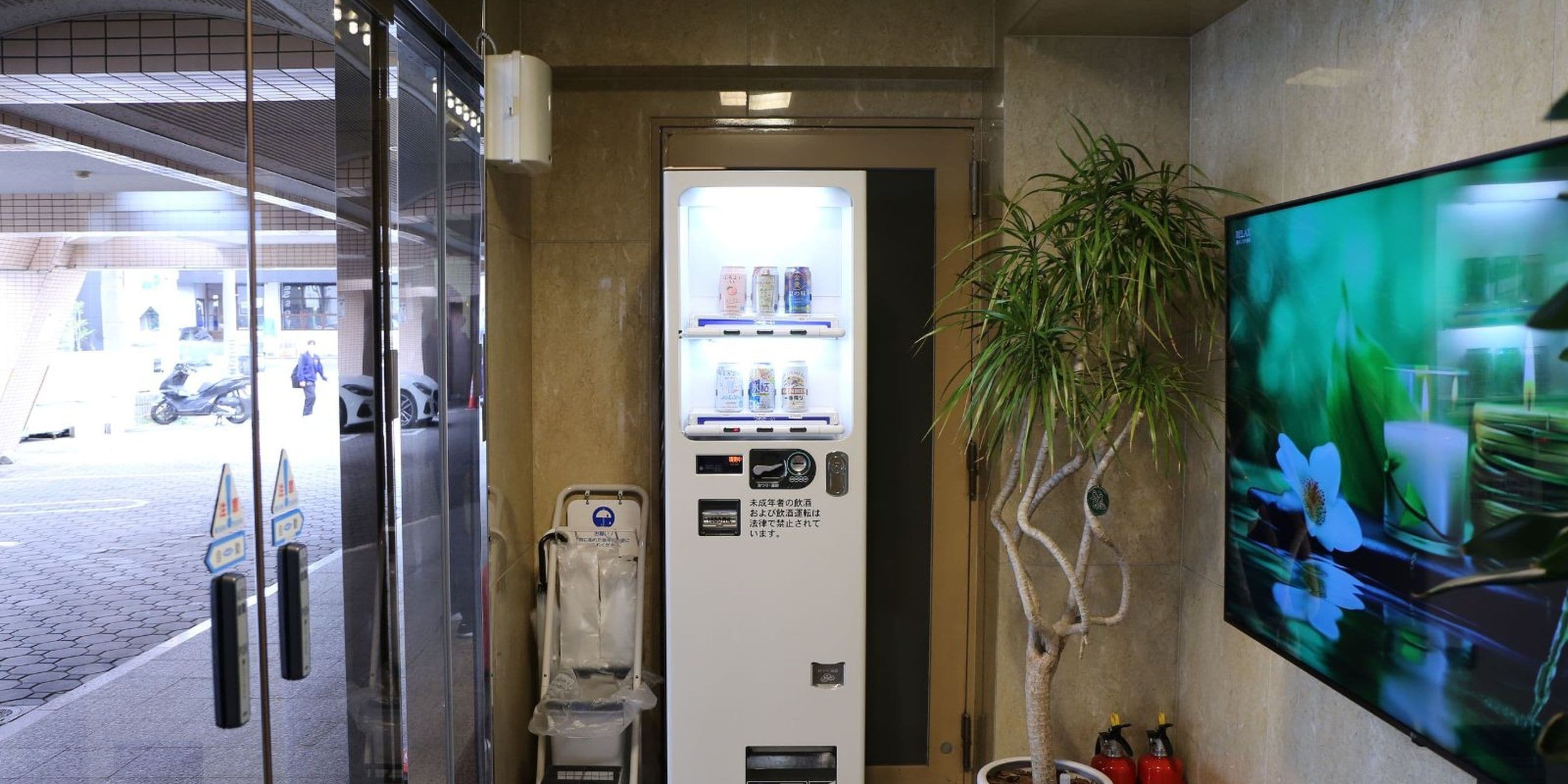 A vending machine with beverages and snacks, a trash bin, and a potted plant.