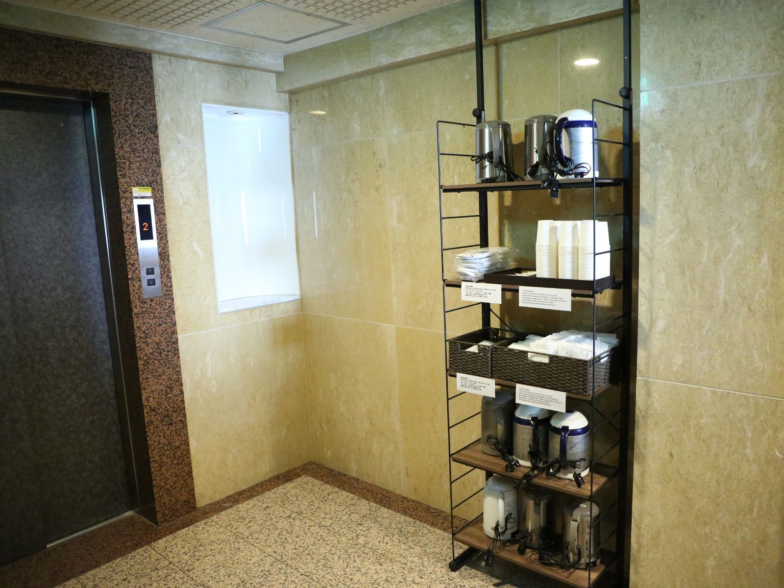 A hotel elevator with a digital display, a metal shelving unit with various items, and a door with a control panel.