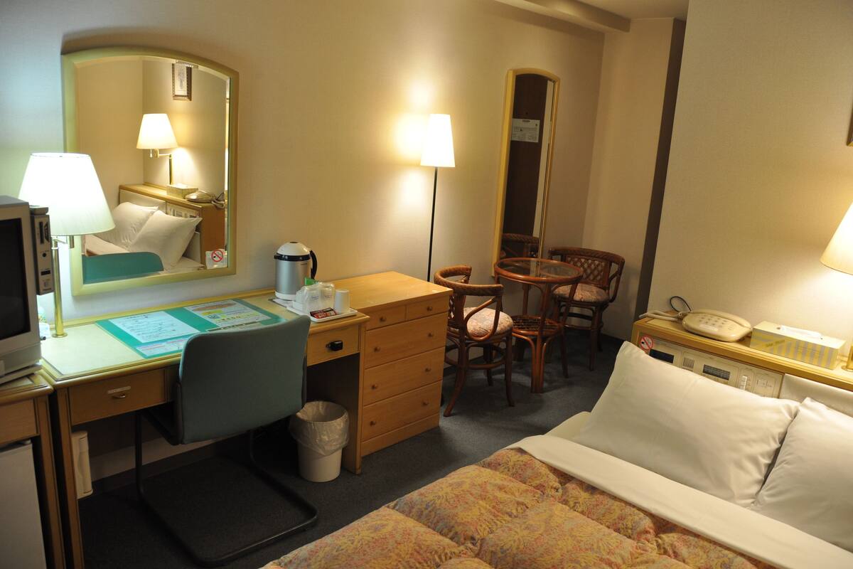 A hotel room with a bed, desk, chair, and a small table with a lamp.