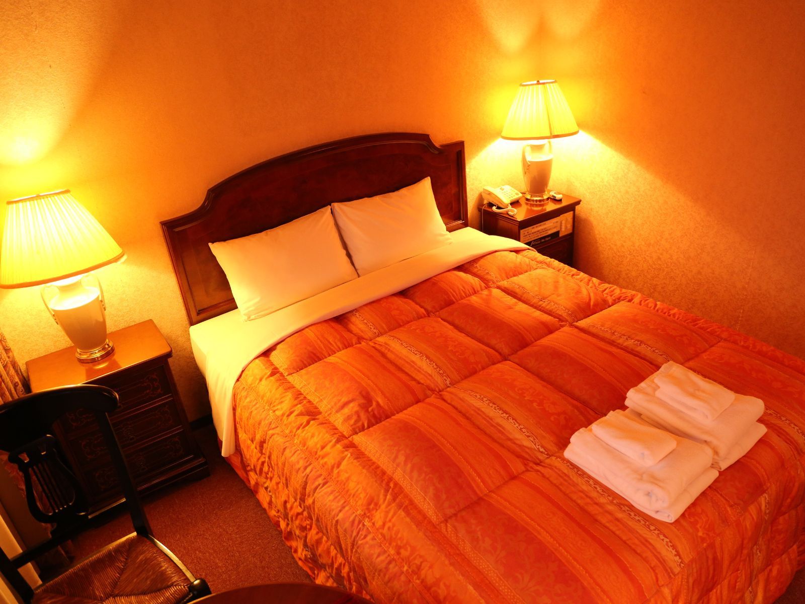 A hotel room with a large bed, two bedside lamps, a chair, and a small table with a lamp.
