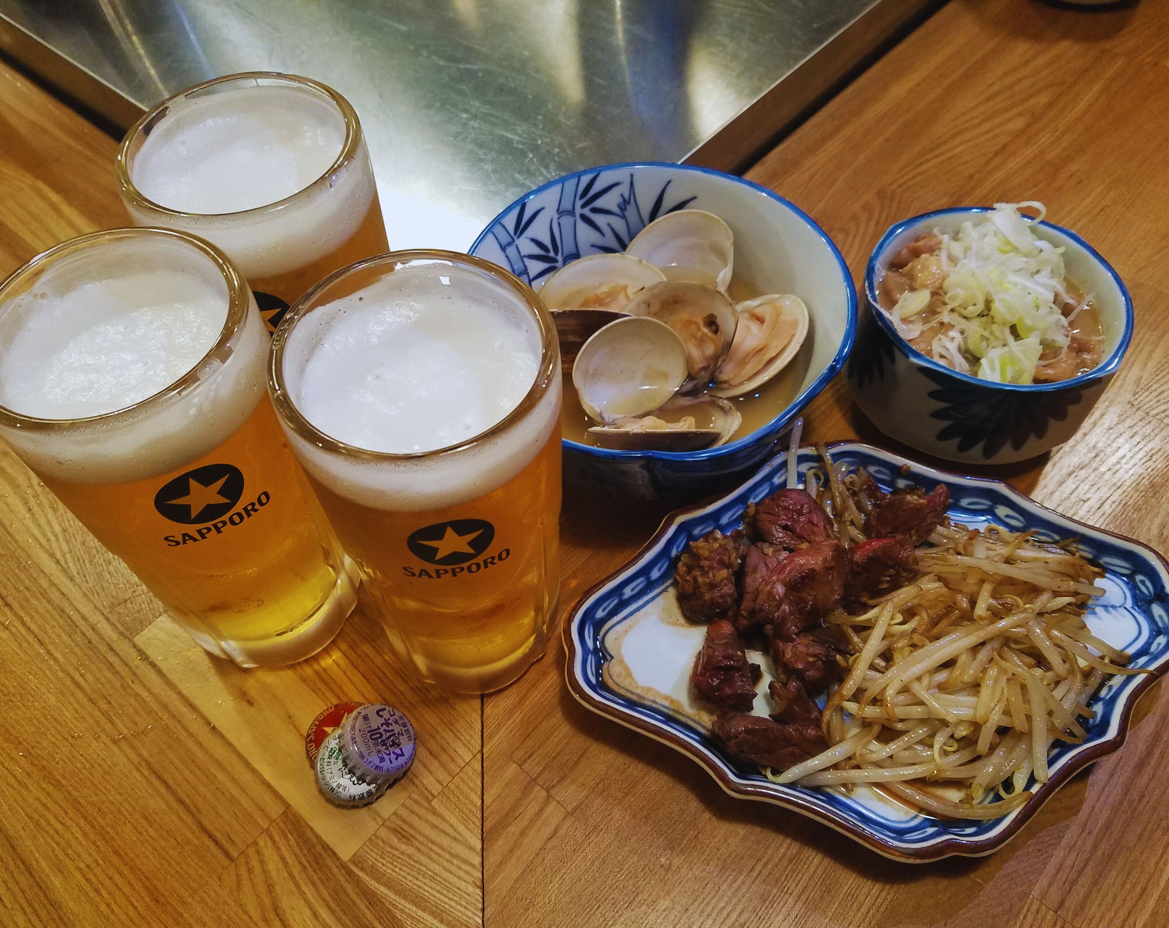 Tres vasos de cerveza, un bol de almejas, un bol de verduras en vinagre y un plato de carne con fideos sobre una mesa de madera.