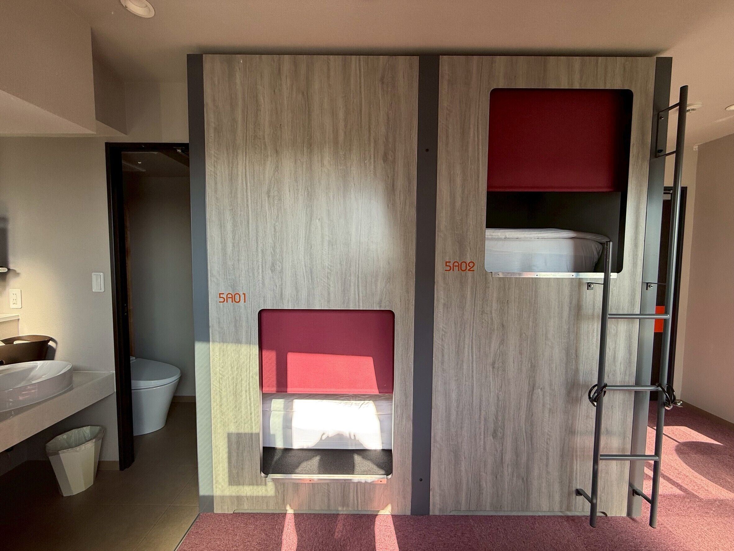 A modern bathroom with a sink, toilet, and two bunk beds with red cushions.