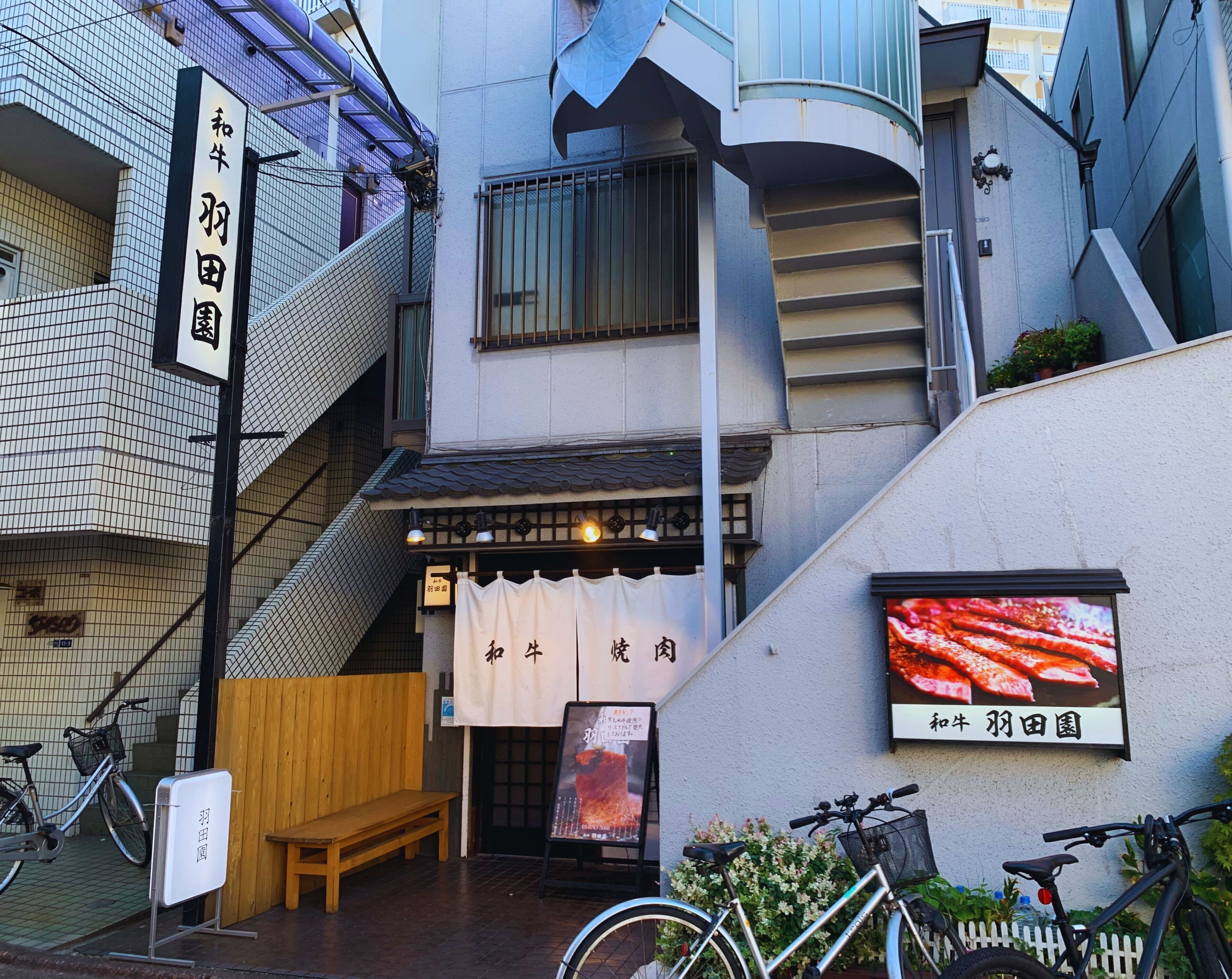 A narrow alley with a restaurant entrance, wooden benches, bicycles, and a signboard.