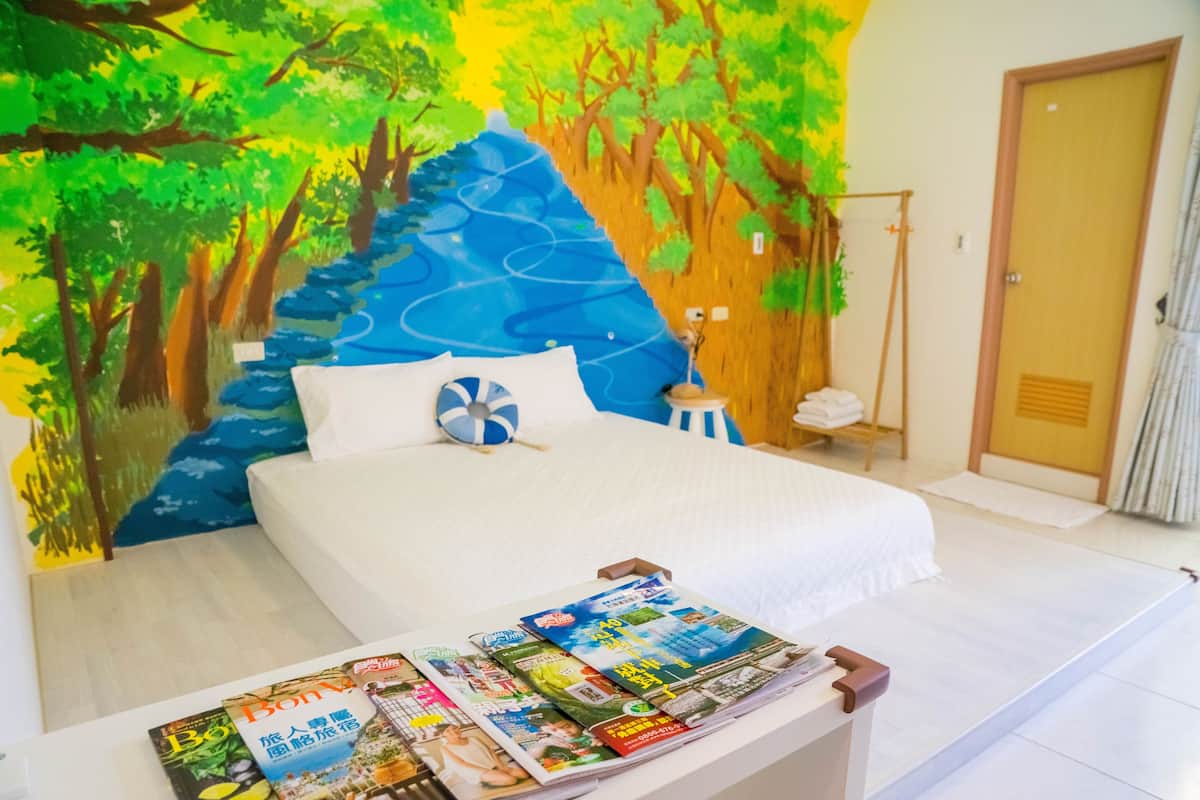A hotel room with a bed, a nightstand, a wall mural, and a magazine rack.