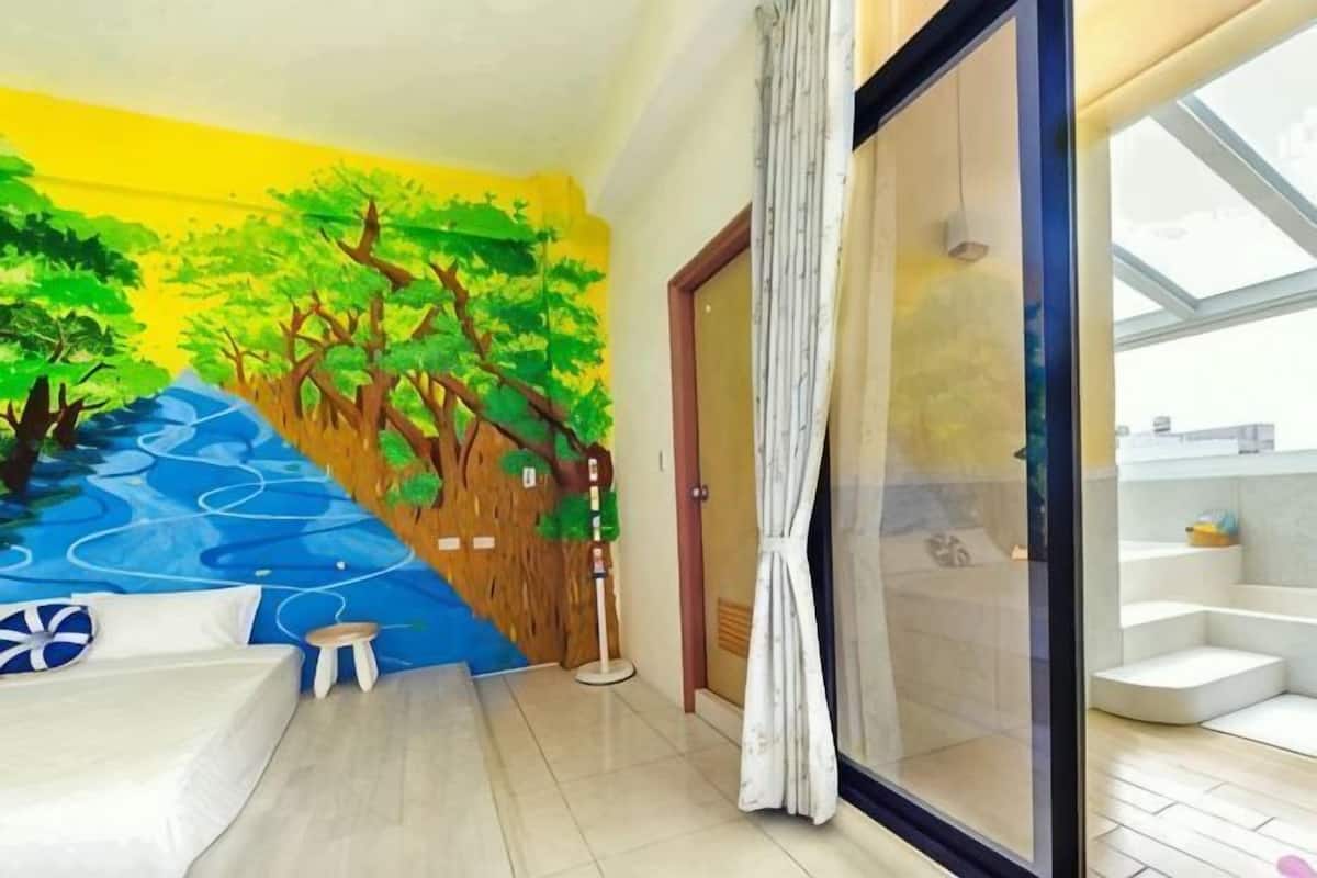 A room with a mural of trees, a bed, a small stool, a sliding glass door, and a view of a bathroom.