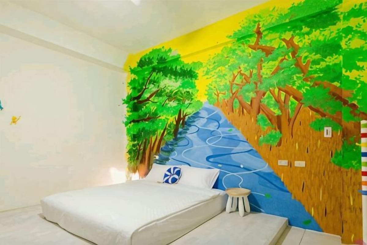 A bedroom with a bed, a small stool, and a wall mural of trees and a river.