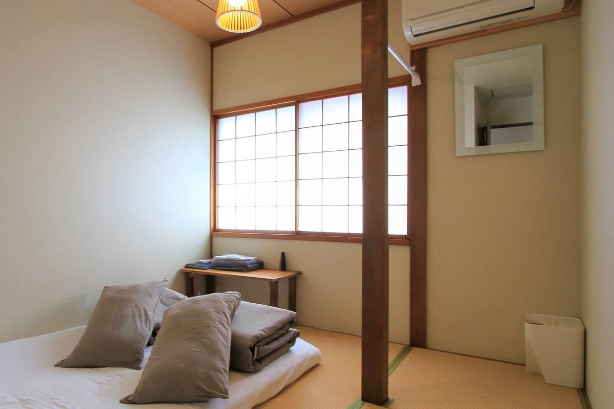 Quarto pequeno e minimalista com tatames, uma cama baixa de madeira com almofadas, uma mesa pequena com abajur e uma janela com vista.