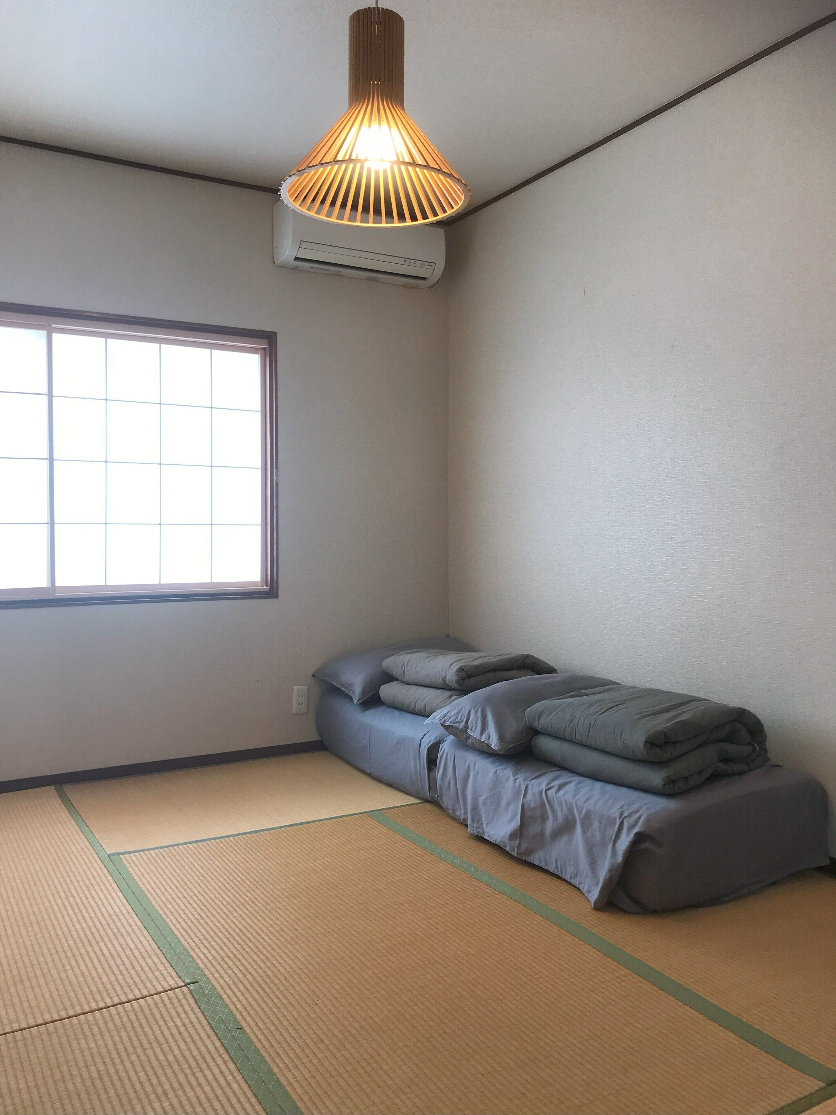 Una habitación pequeña y poco amueblada con suelo de esteras de tatami, una cama individual con ropa de cama gris, una ventana que proporciona luz natural y una lámpara colgante.