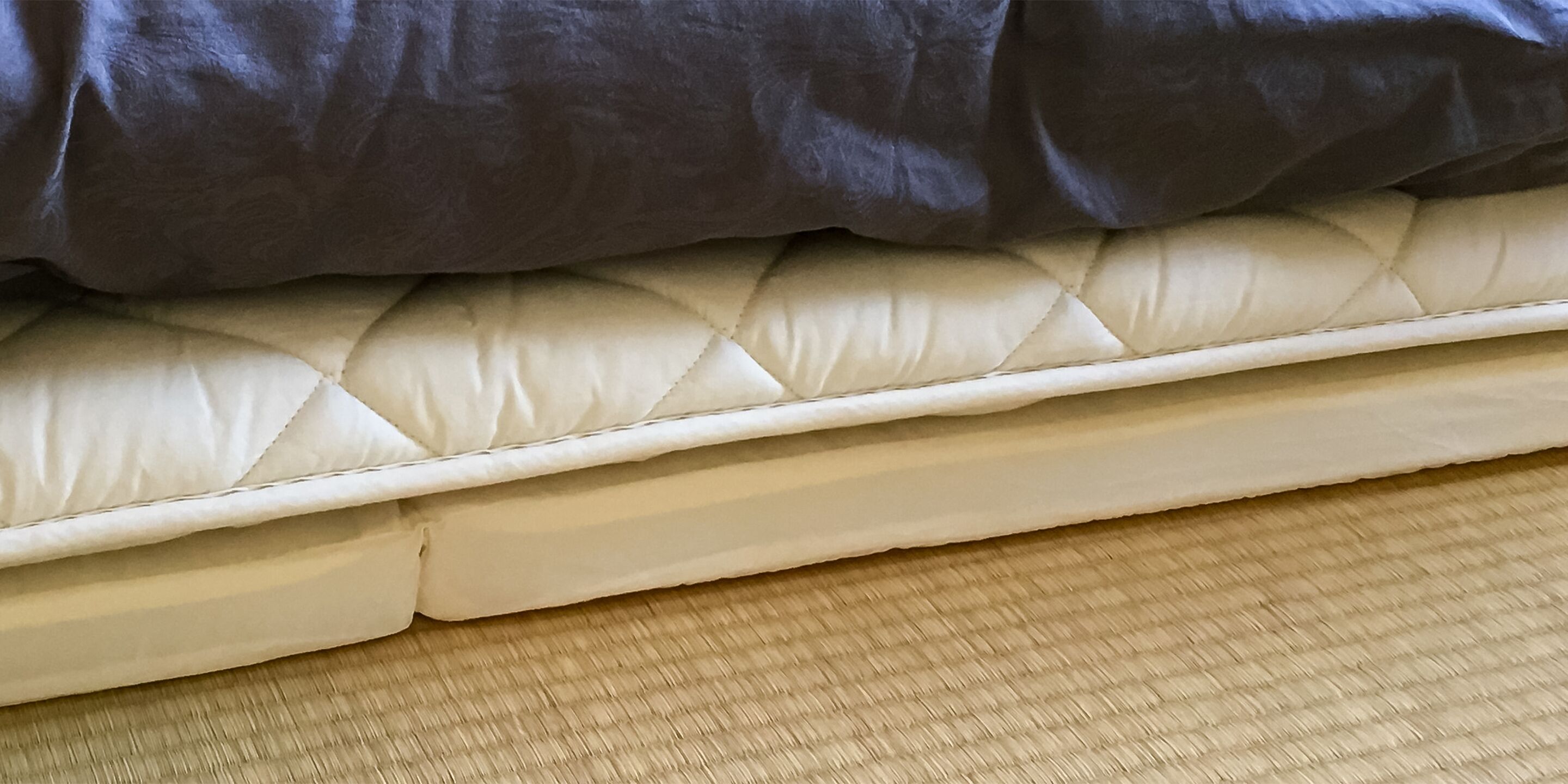 A bed with a quilted white mattress and a dark grey blanket.