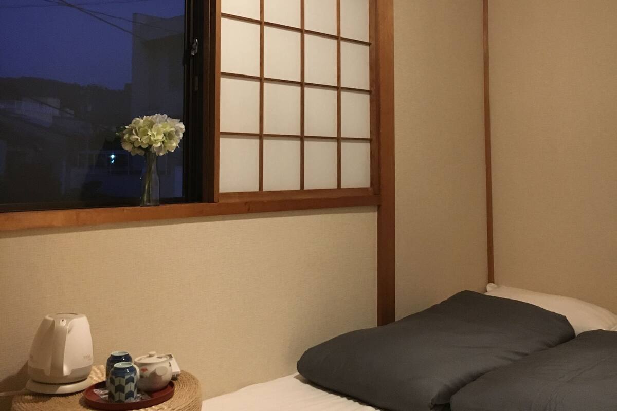 Um quarto em estilo japonês tradicional com tatames, uma cama baixa com almofadas escuras, uma pequena mesa com um vaso e bule, e um painel shoji sobre uma janela.