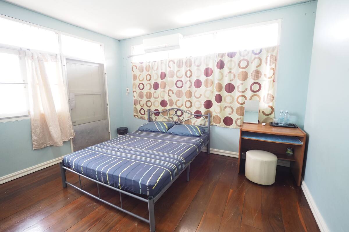 A bedroom with a bed, a desk, a chair, a small stool, a window with curtains, and a door.