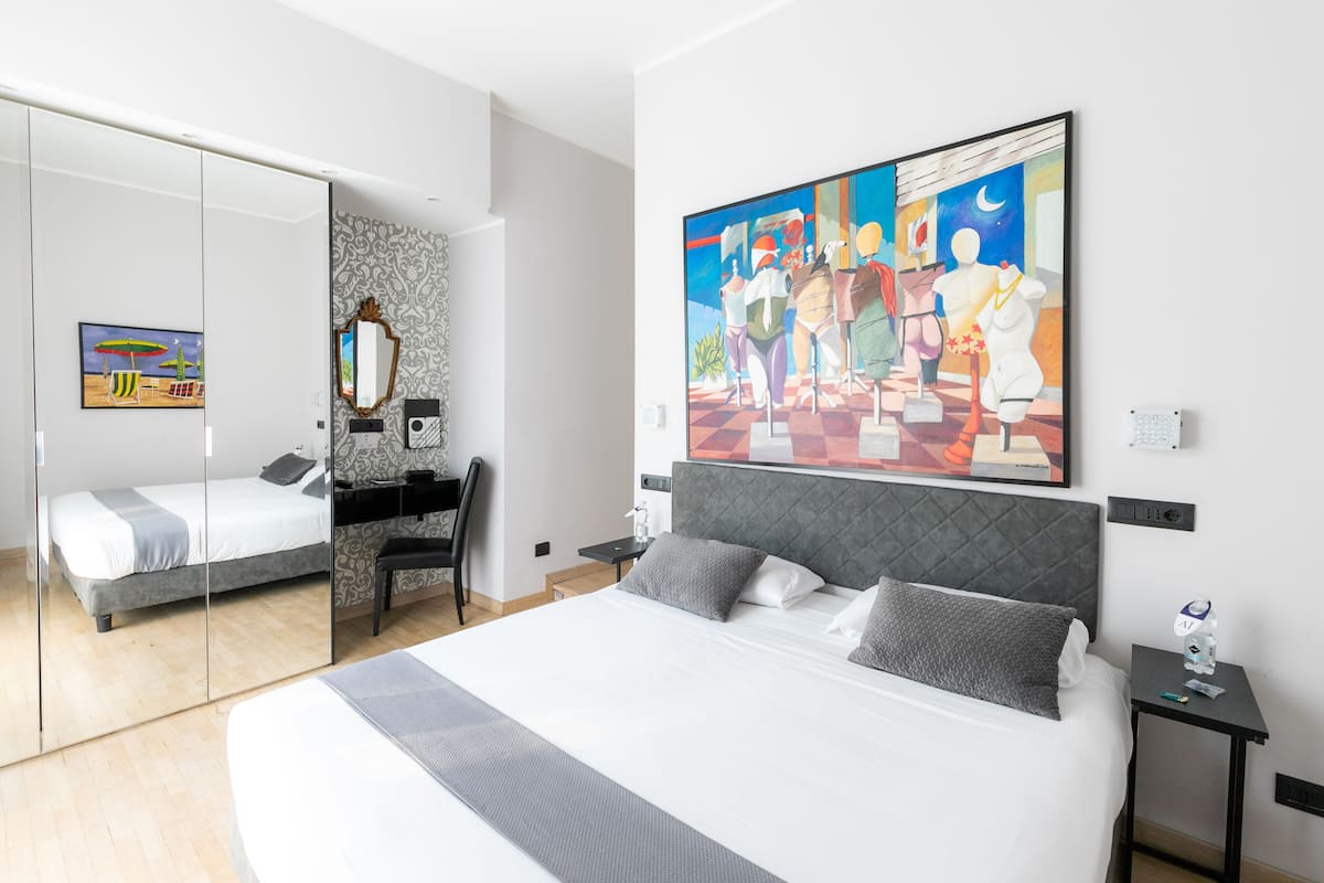A modern hotel room with a large bed, a desk, and a chair. There is a painting on the wall and a mirror on the door.