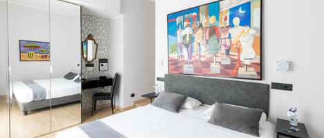 A modern hotel room with a large bed, a desk, and a chair. There is a painting on the wall and a mirror on the door.